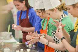 A team of young people wearing chefs hats and apron, rolling some dough on a kitchen counter top surface