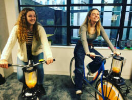 Two smiling young ladies on blender bikes side by side, blending orange juice