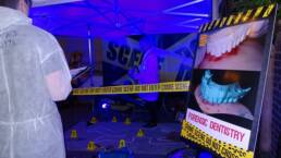 Forensic Dentistry board surrounded by tape which reads crime scene - do not cross. A bystander wearing a CSI suit is observing the scene