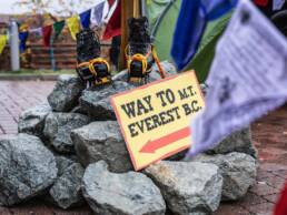 A pair of climbing boots on a pile of rocks with a 'Way to Everest' sign to guide teams towards the event. Prayer flags surround it