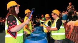 A team of six people wearing hard hats and high visibility vests using barrels as drums in team-building event Junk Funk