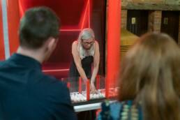 A lady is in a qube playing seperate, a challenge involving separating white and red balls. Two of her colleges are watching her