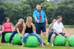 A team of adults are sat on space hoppers whilst a marshal is instructing them, getting them ready to race