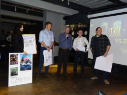 Five people holding signs pitching Dragons Den with a powerpoint presentation in the background, with the dragons den title screen displayed as part of the Dragons Den team-building event